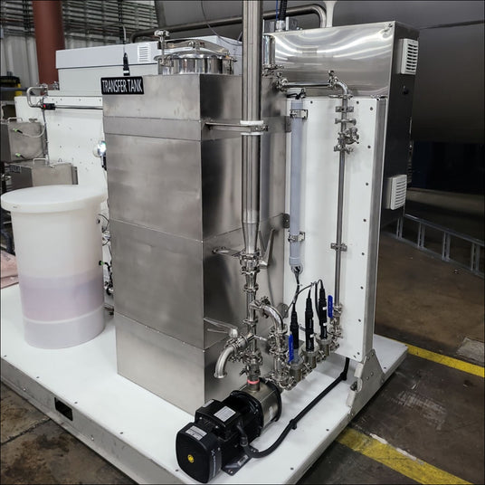 Stainless steel transfer tank with connected pipes and a motor, set in a warehouse.