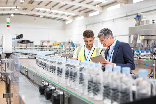 Two men discussing in a factory setting, with a production line of water bottles in the background.
