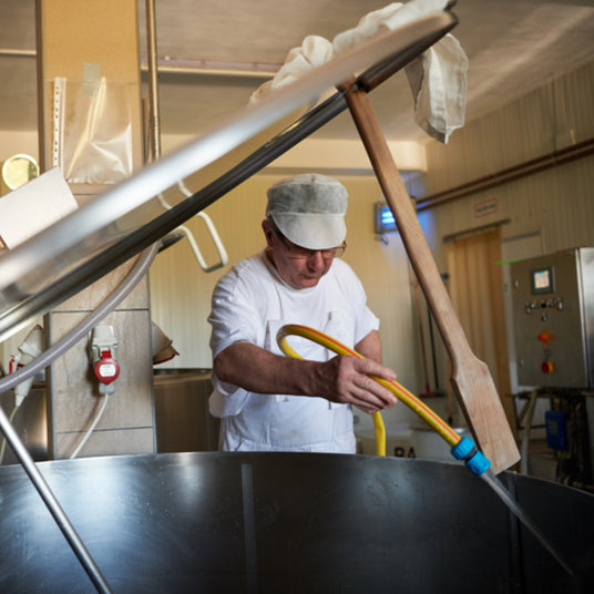 Man in a white shirt and apron using a hose and wooden paddle in a large metal container.