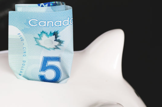 Five Canadian dollar bill partially inserted into a white piggy bank.