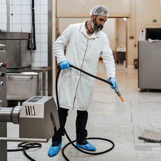 A man in a white lab coat and hairnet cleaning a surface with a pressure washer in a kitchen environment.
