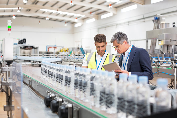 Two men discussing in a factory setting, with a production line of water bottles in the background.