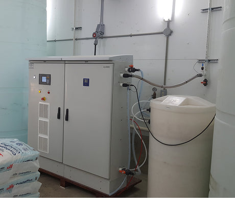 Industrial equipment with a control panel, connected to a large white tank and bags of material in a light-colored room.