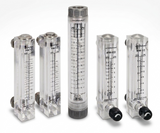 Five clear plastic flow meters with black measurement markings and black fittings, arranged on a light gray studio background. The set includes different flow meter models, with one central taller meter and four shorter side meters.