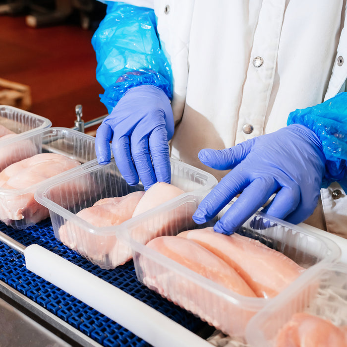 Hands in blue gloves handling packaged raw chicken fillets in a food processing setting.