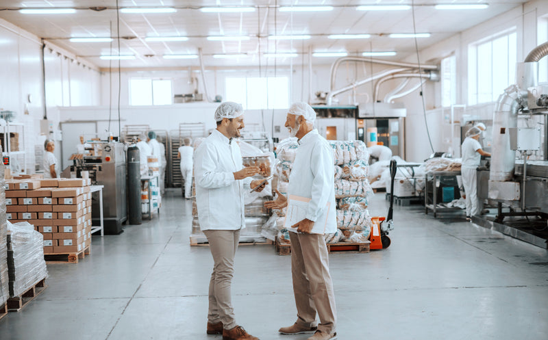 Two men in white lab coats and hairnets discussing in a food processing facility with boxes and bags in the background.