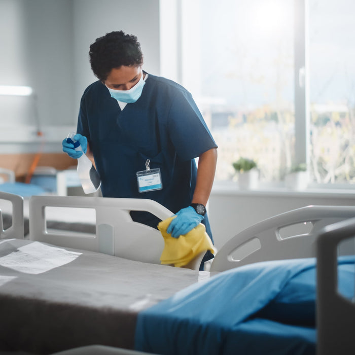 Healthcare worker cleaning a hospital bed with a yellow cloth and spray bottle.
