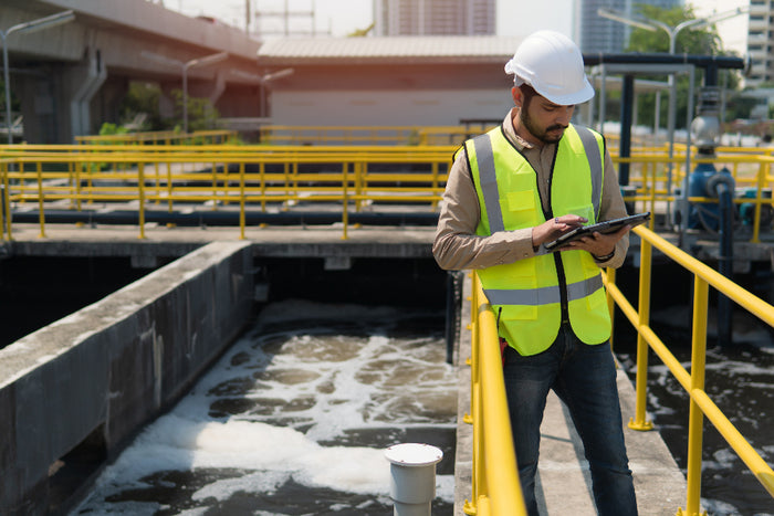 Man in a safety vest and hard hat using a tablet while standing near a water treatment facility with yellow railings.