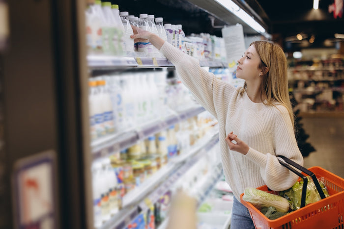 Woman shopping for drinks in a grocery store aisle, reaching for a bottle on the shelf while holding a shopping basket with vegetables.